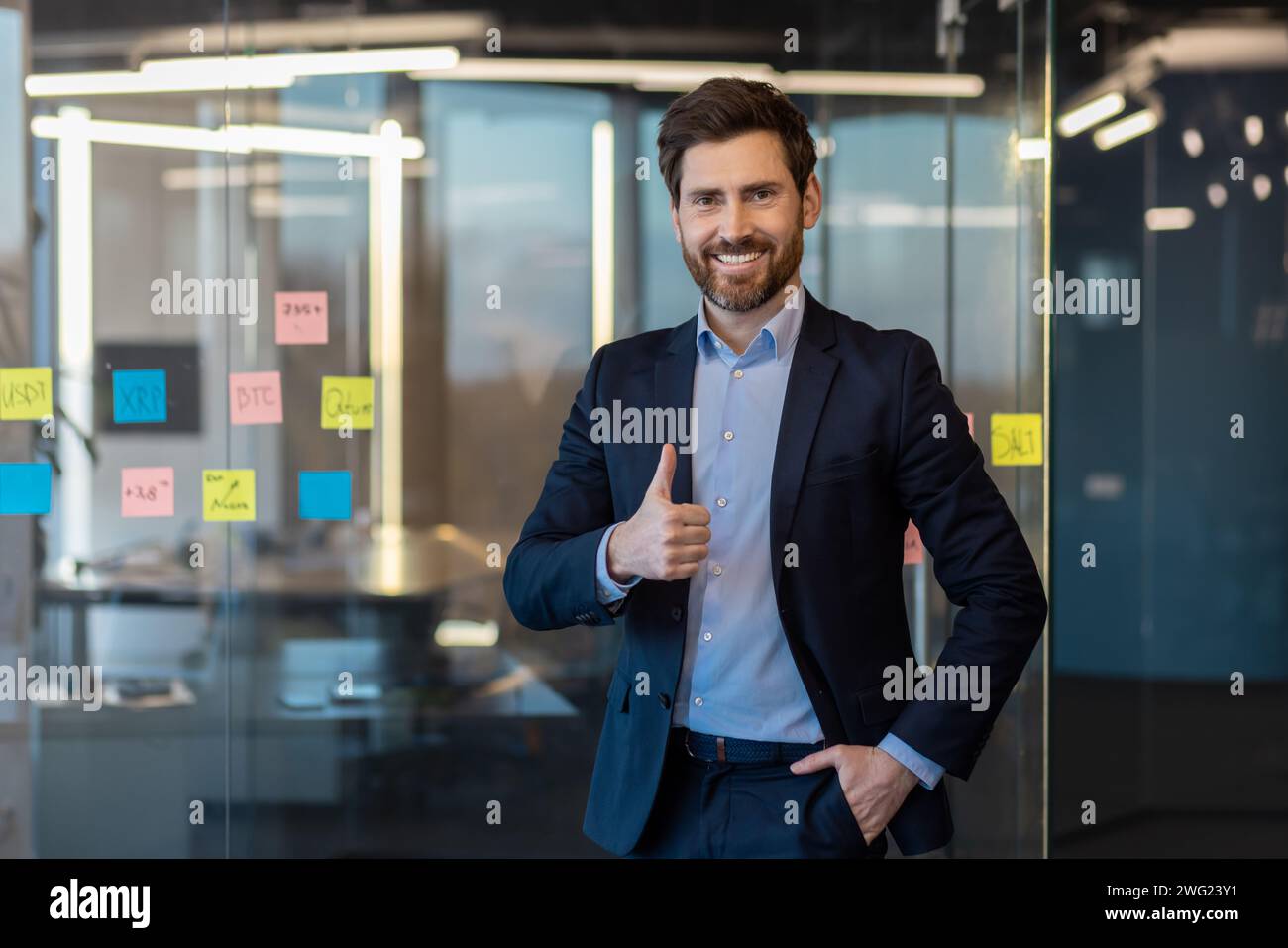Erfolgreicher und lächelnder junger Geschäftsmann, der in die Kamera blickt und ein Superzeichen mit dem Finger zeigt und die Hand in der Tasche hält. Selbstbewusst in einem Anzug im Büro stehen. Stockfoto