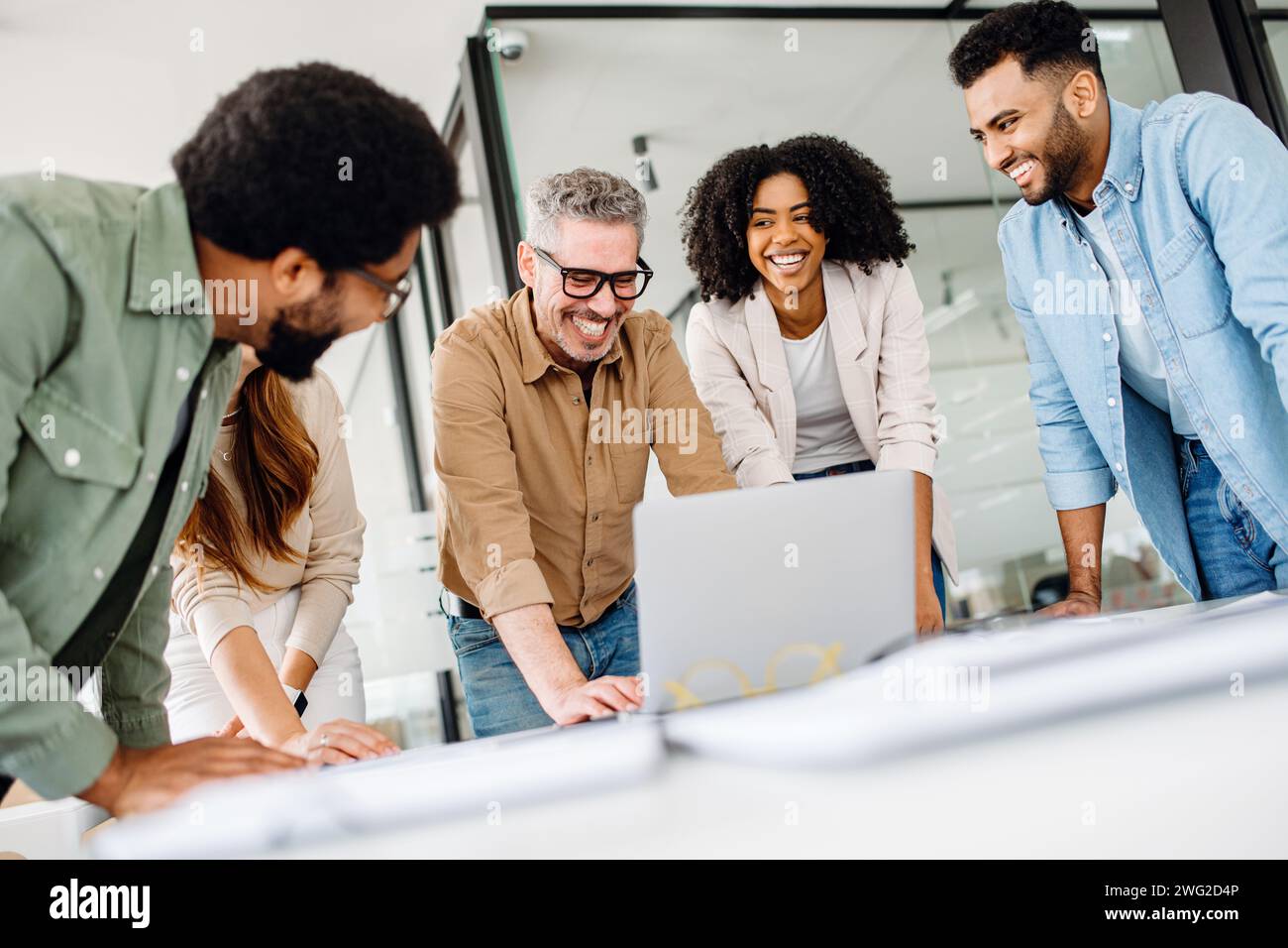 Eine fröhliche Gruppe verschiedener Kollegen lehnt sich um einen Laptop herum, was auf eine interaktive Diskussion über die Technik und die Stimmung eines produktiven und zukunftsorientierten Teams hindeutet. Stockfoto