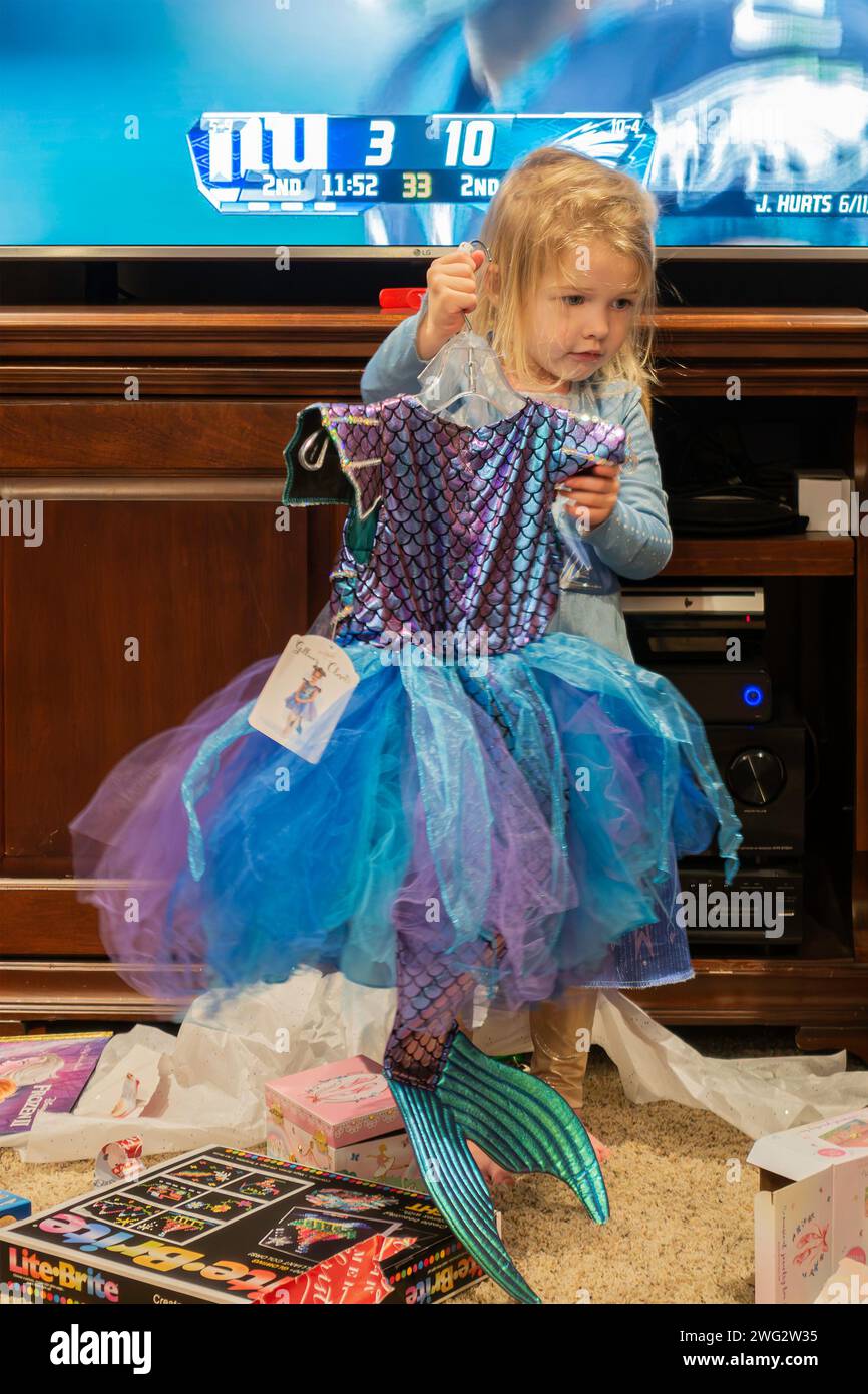 Ein dreijähriges blondes weißes Mädchen, das ein Meerjungfrauenkostüm hochhält, das sie als Geschenk erhalten hat. USA. Stockfoto