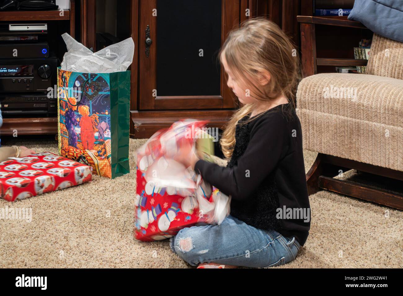 Ein 5-6-jähriges weißes Mädchen sitzt auf dem Boden und reißt am Weihnachtstag Geschenkverpackung von einem Weihnachtsgeschenk ab. USA. Stockfoto