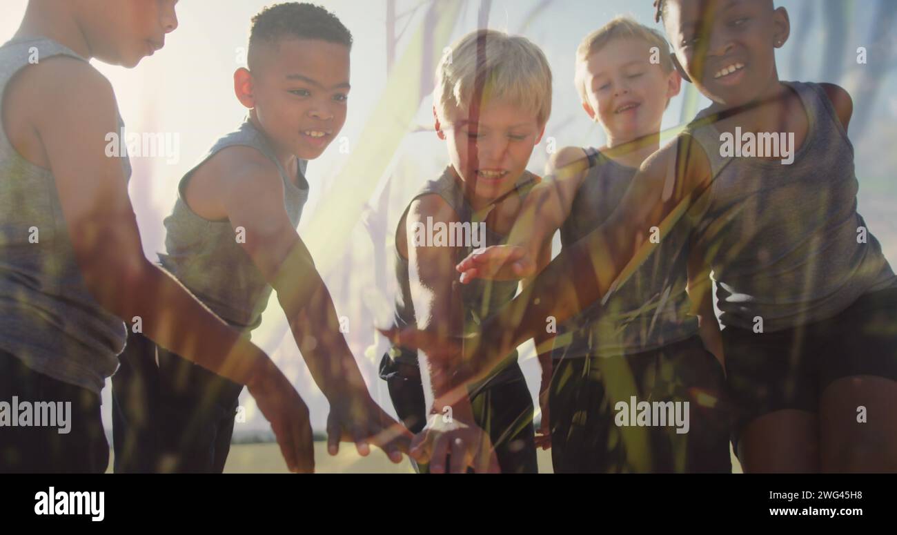 Bild von Sonne und Gräsern über glücklichen, vielfältigen Schulkindern, die Hände in Outdoor-Sportklassen stapeln Stockfoto