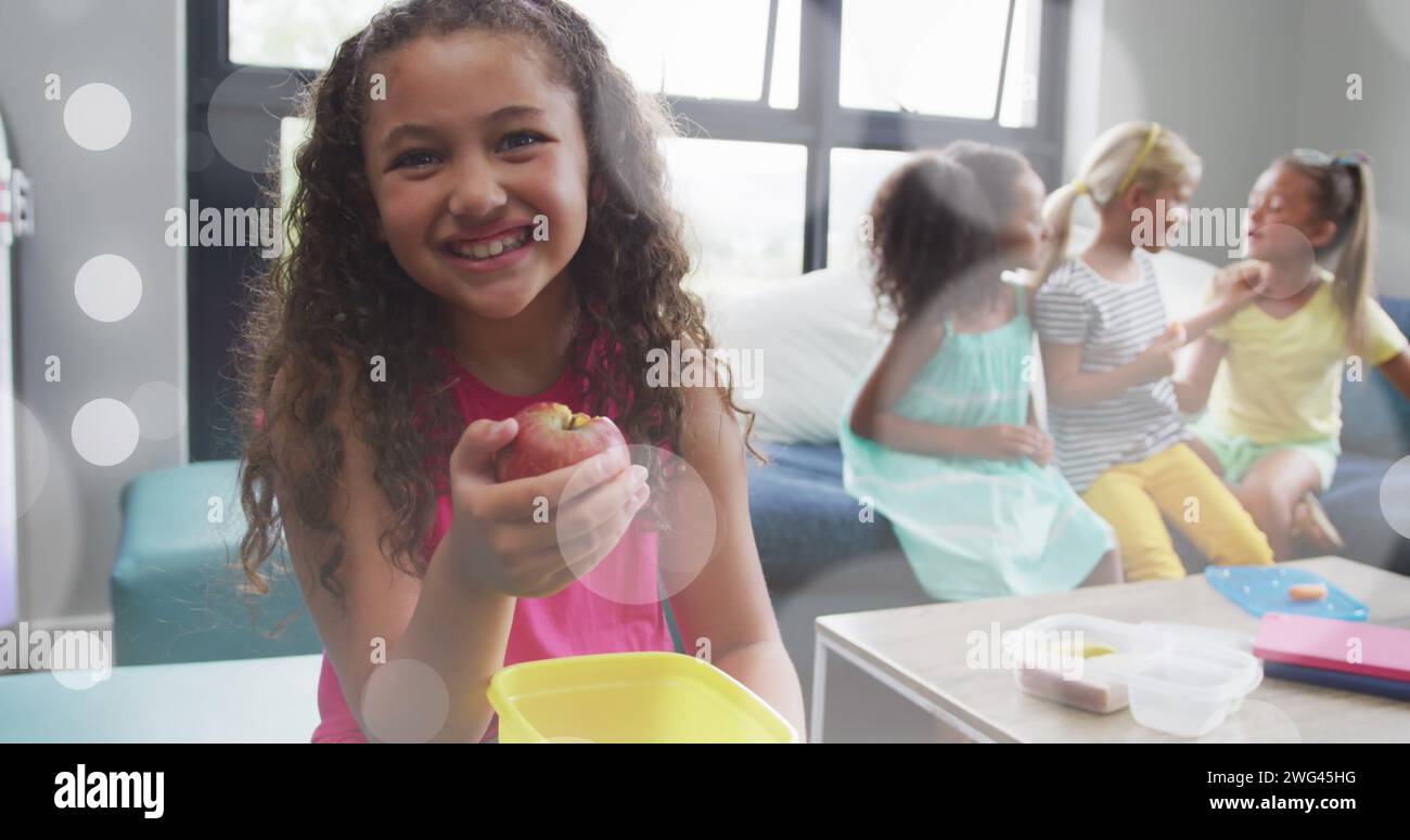 Bild von weißen Bokeh-Lichtflecken über glücklichen verschiedenen Schulmädchen, die Lunchpakete essen und reden Stockfoto