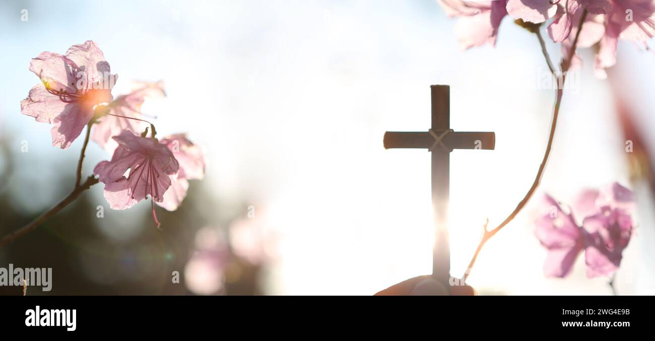 Hintergrund mit starkem Sonnenlicht, das an einem Frühlingstag durch Azaleen-Blüten leuchtet, und einem Christen, der das heilige Kreuz Jesu Christi in seinen Händen hält Stockfoto