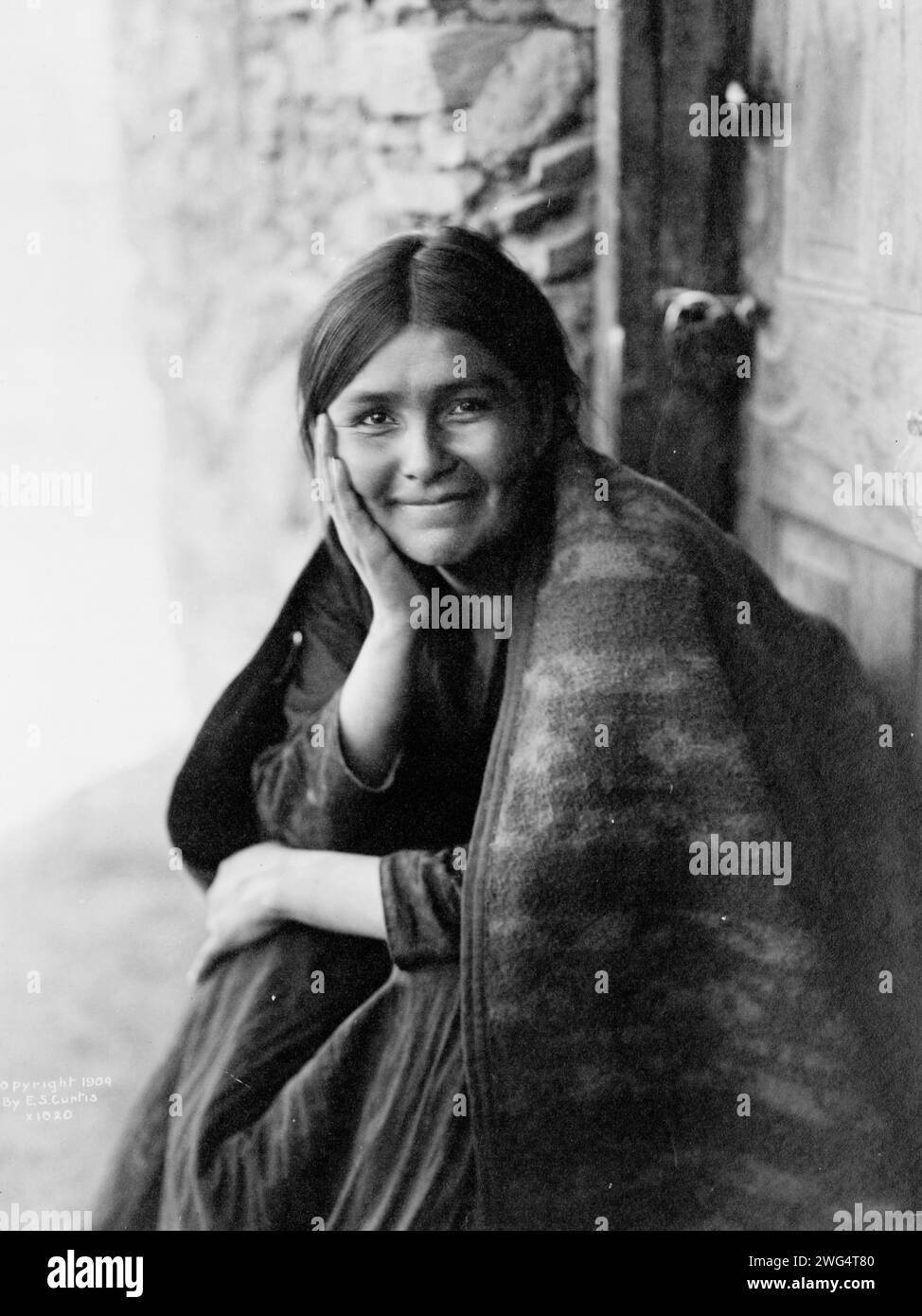 Ein Navaho-Lächeln, 1904. Das Foto zeigt eine Navaho-Frau, die vor der Tür sitzt, mit der rechten Hand auf der Wange, nach vorne gerichtet, lächelnd. Stockfoto