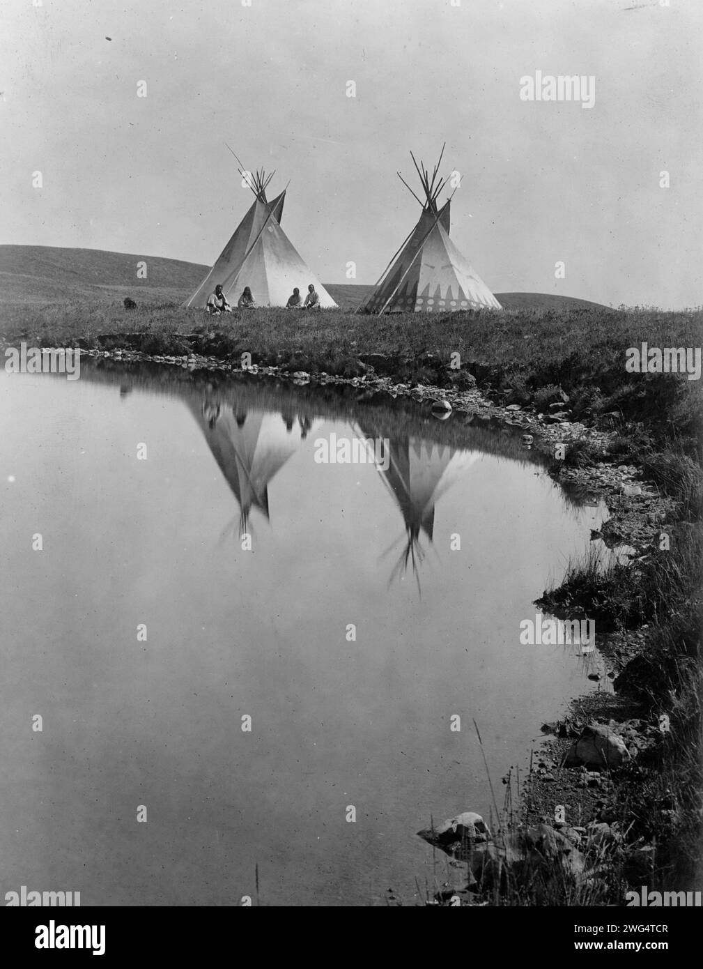 Am Wasserrand - Piegan, 1910. Das Foto zeigt zwei Tipis, die sich im Wasser des Teichs spiegeln, mit vier Piegan-Indianern, die vor einem Tipi sitzen. Stockfoto