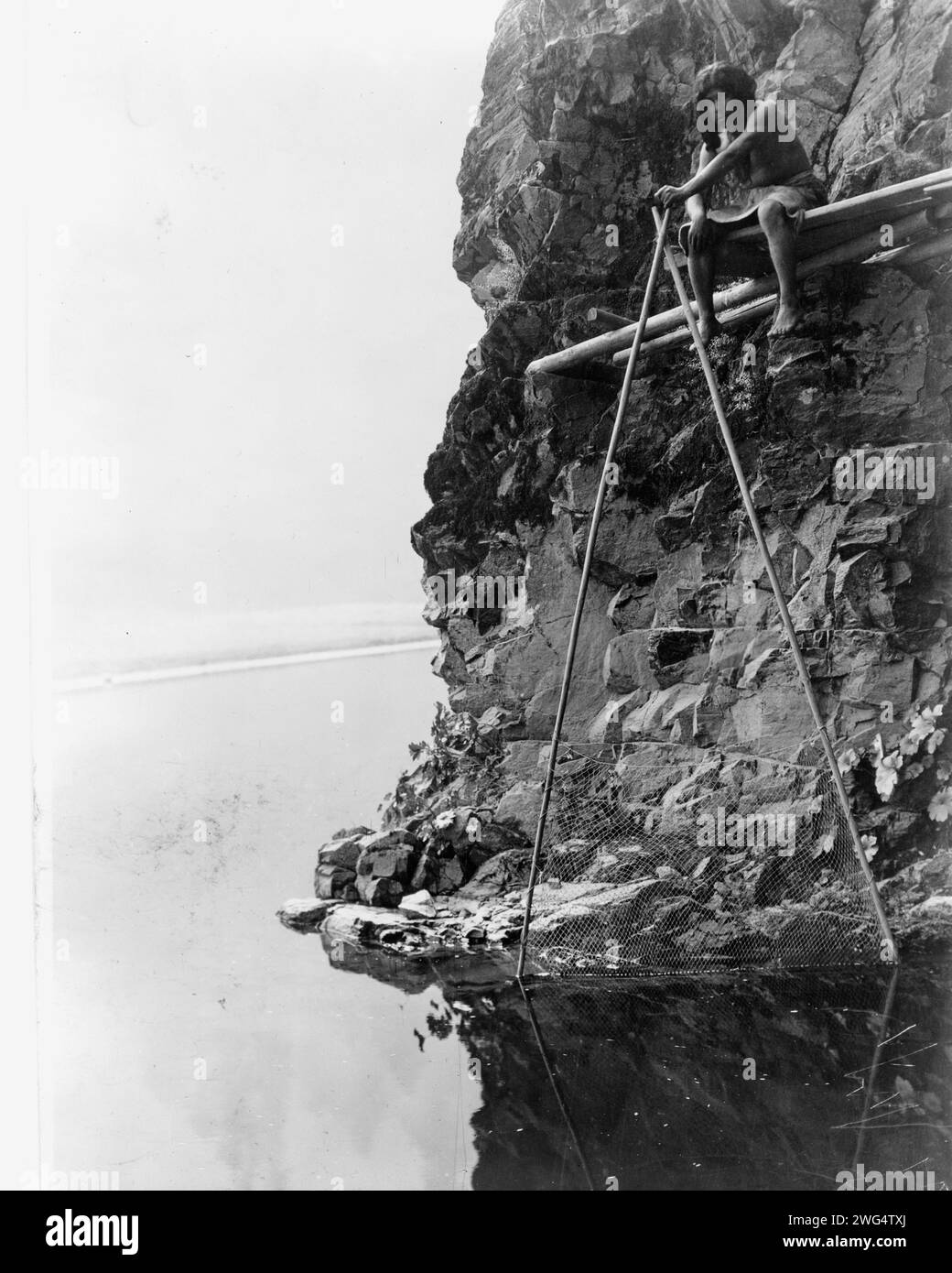 Angelplattform am Trinity River-Hupa, um 1923. Das Foto zeigt einen Hupa-Mann, der auf einer Plattform auf einer felsigen Klippe sitzt und mit einem Fischernetz umgeht. Stockfoto