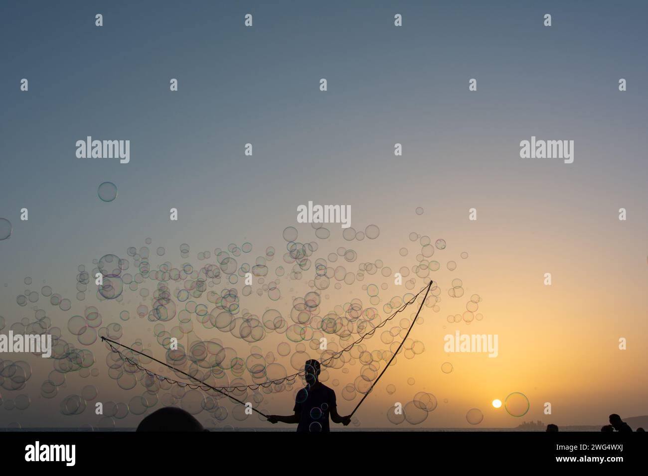 Ein Mann in der Silhouette macht viele Seifenblasen mit einem Seifenblasenstab, sie schweben am orangefarbenen Himmel bei Sonnenuntergang Stockfoto