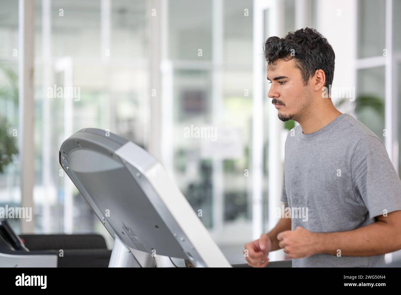 Das Engagement eines Mannes für seine Fitnessziele wird deutlich, wenn er sich durch ein Laufband-Workout in einem sonnendurchfluteten Fitnessstudio anstrengt Stockfoto