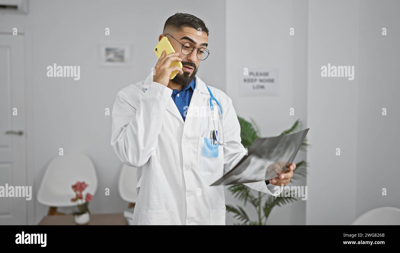 Ein bärtiger Mann in weißem Mantel untersucht Röntgenaufnahmen, während er im Wartezimmer einer Klinik telefoniert. Stockfoto