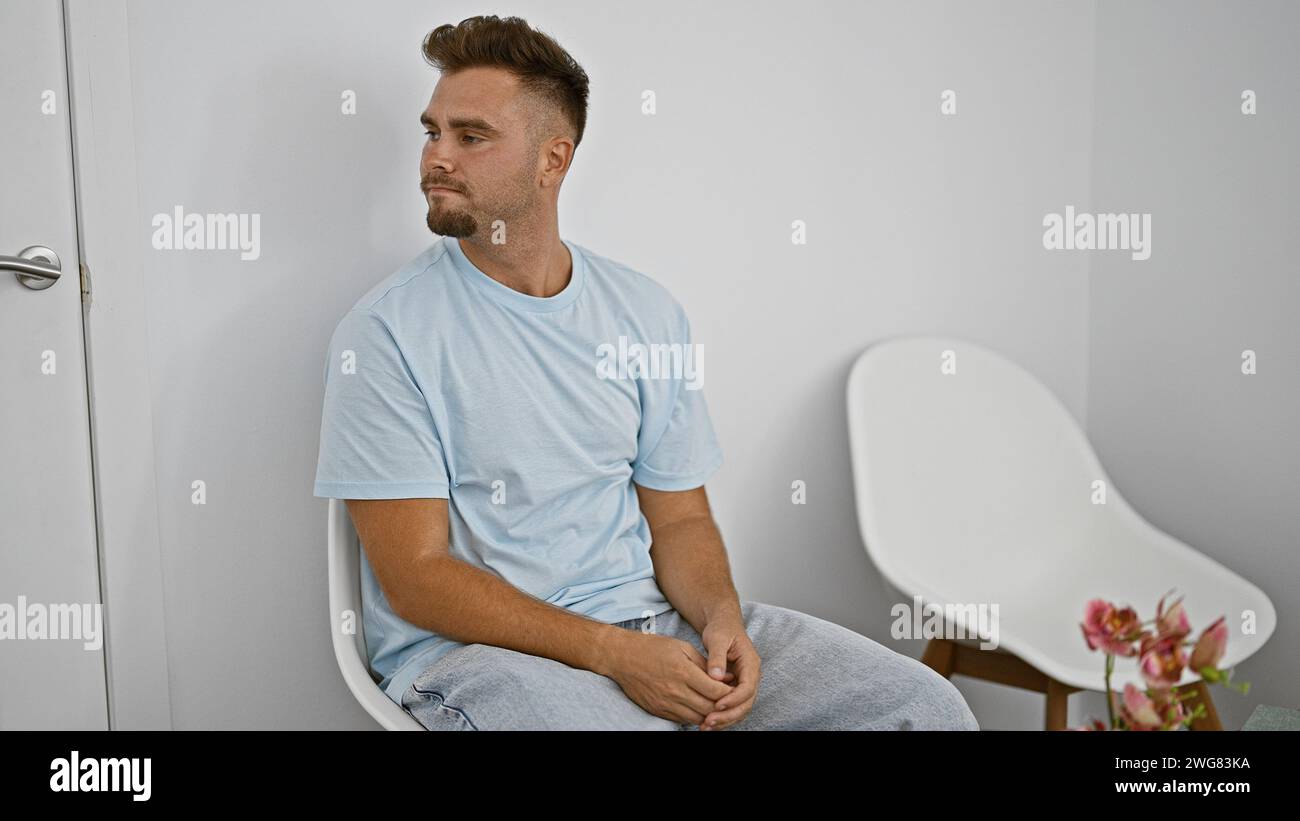 Ein nachdenklicher junger Mann mit Bart sitzt geduldig in einem minimalistischen weißen Raum und spiegelt einen Hauch der Ruhe wider. Stockfoto