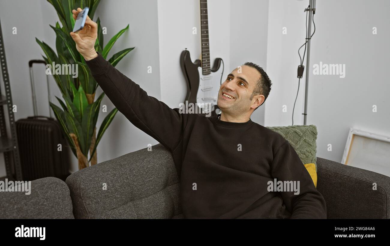 Ein fröhlicher junger Mann nimmt ein Selfie auf seinem Handy auf, während er bequem auf einer Couch in einem modernen Wohnzimmer sitzt. Stockfoto