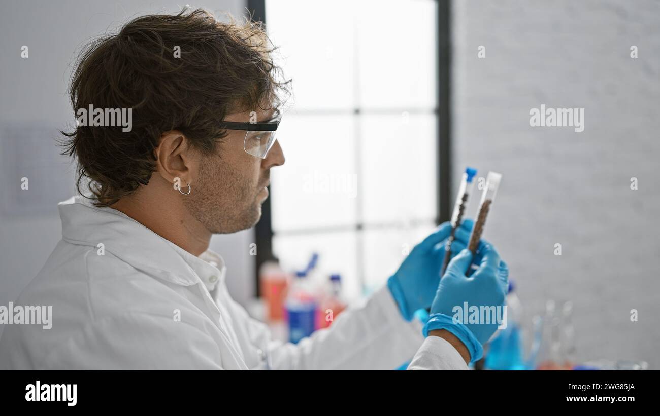 Ein bärtiger Mann in Schutzbrille und -Handschuhen untersucht Reagenzgläser in einem hellen Labor. Stockfoto
