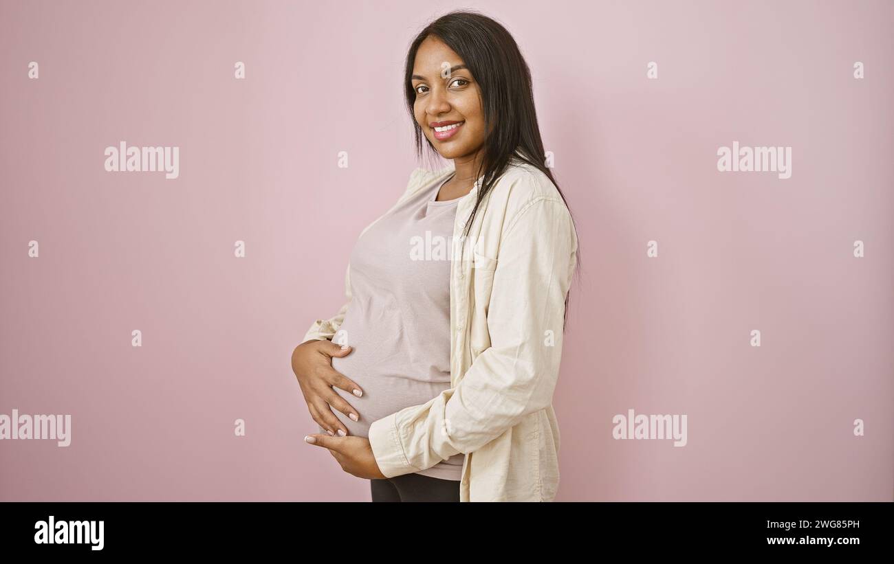 Fröhliche junge schwangere Frau mit einem strahlenden Lächeln berührt freudig ihren Bauch und steht beiläufig über rosa isoliertem Hintergrund Stockfoto