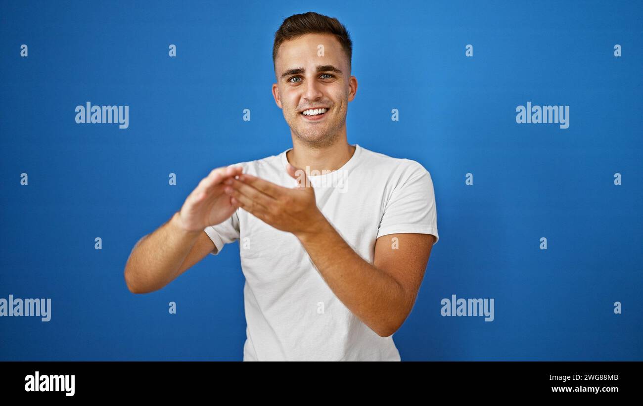 Ein lächelnder junger hispanischer Mann in einem weißen Hemd zeigt vor einem isolierten blauen Hintergrund Gesten, die Positivität und Attraktivität verkörpern. Stockfoto
