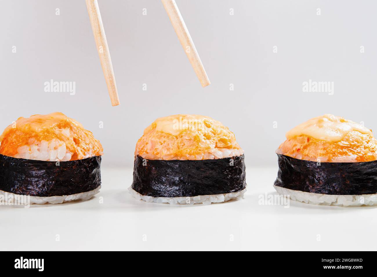 Ein Hauch von Essstäbchen, die positioniert werden, um ein Stück aus einem Trio von scharfen gebackenen Sushi-Nigiri oder Lachsgunkans auf weißem Hintergrund auszuwählen Stockfoto