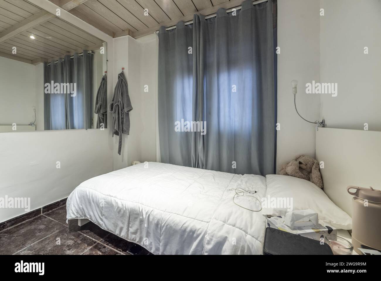 Ein kleines Schlafzimmer mit weißen Wänden und Holzkassettendecke, ein Einzelbett mit weißer Federdecke, graue Vorhänge am Fenster und große Spiegel Stockfoto