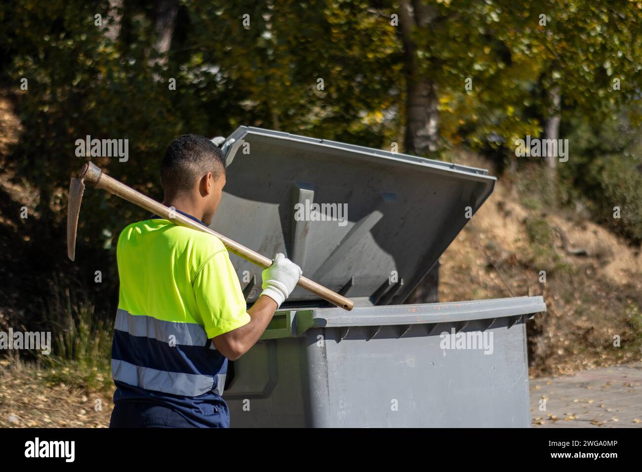 Rückansicht eines lateinischen kommunalen Müllarbeiters in Uniform, der mit einer Sichel über der Schulter Müll in den Müllcontainer wirft Stockfoto