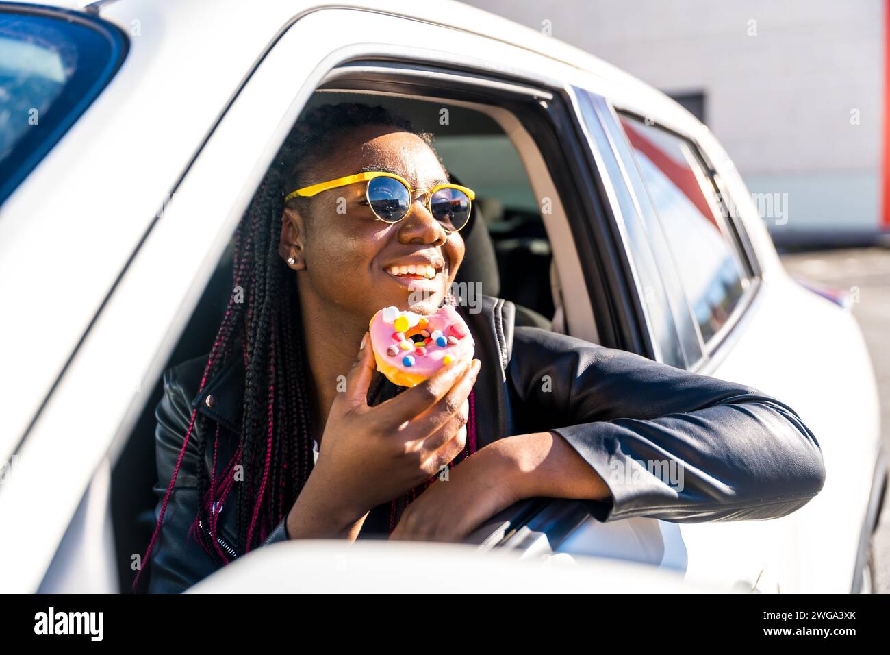 Eine abgelenkte Frau isst Süßigkeiten in einem Auto mit Arm, der auf das Fenster gebeugt ist, und blickt auf die urbane Landschaft Stockfoto