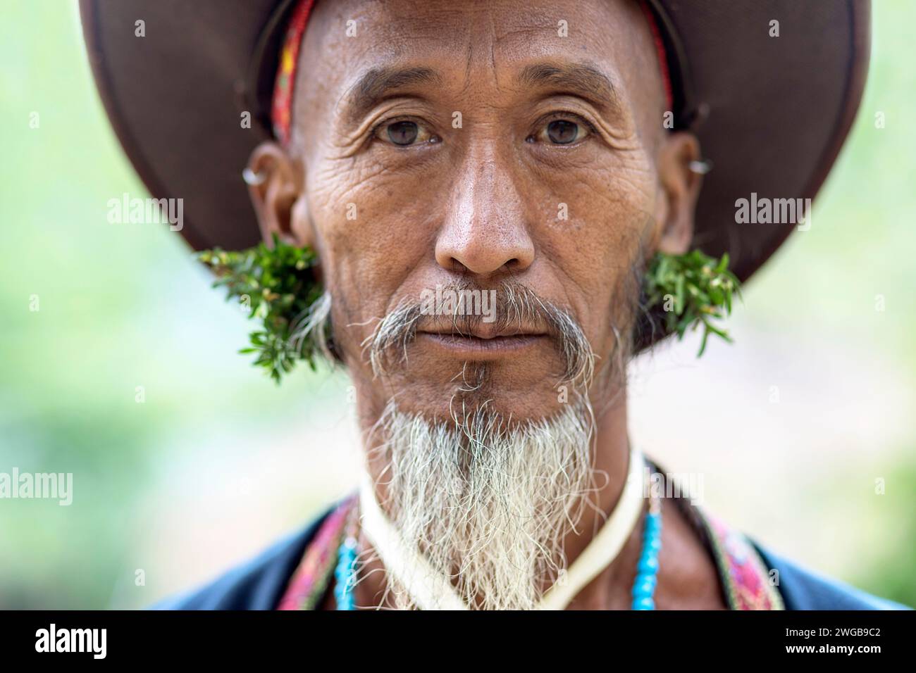 Porträt eines Mannes aus dem Stamm der Wancho Naga in seinen traditionellen Kleidern mit einem traditionellen Dorf im Hintergrund, Arunachal pradesh, Indien Stockfoto