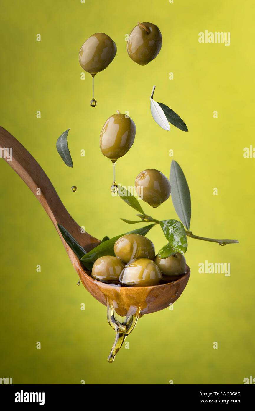 Kelle mit Oliven und Öl auf grünem Hintergrund. Oliven, natives Olivenöl extra und Olivenblätter schweben in der Luft Stockfoto