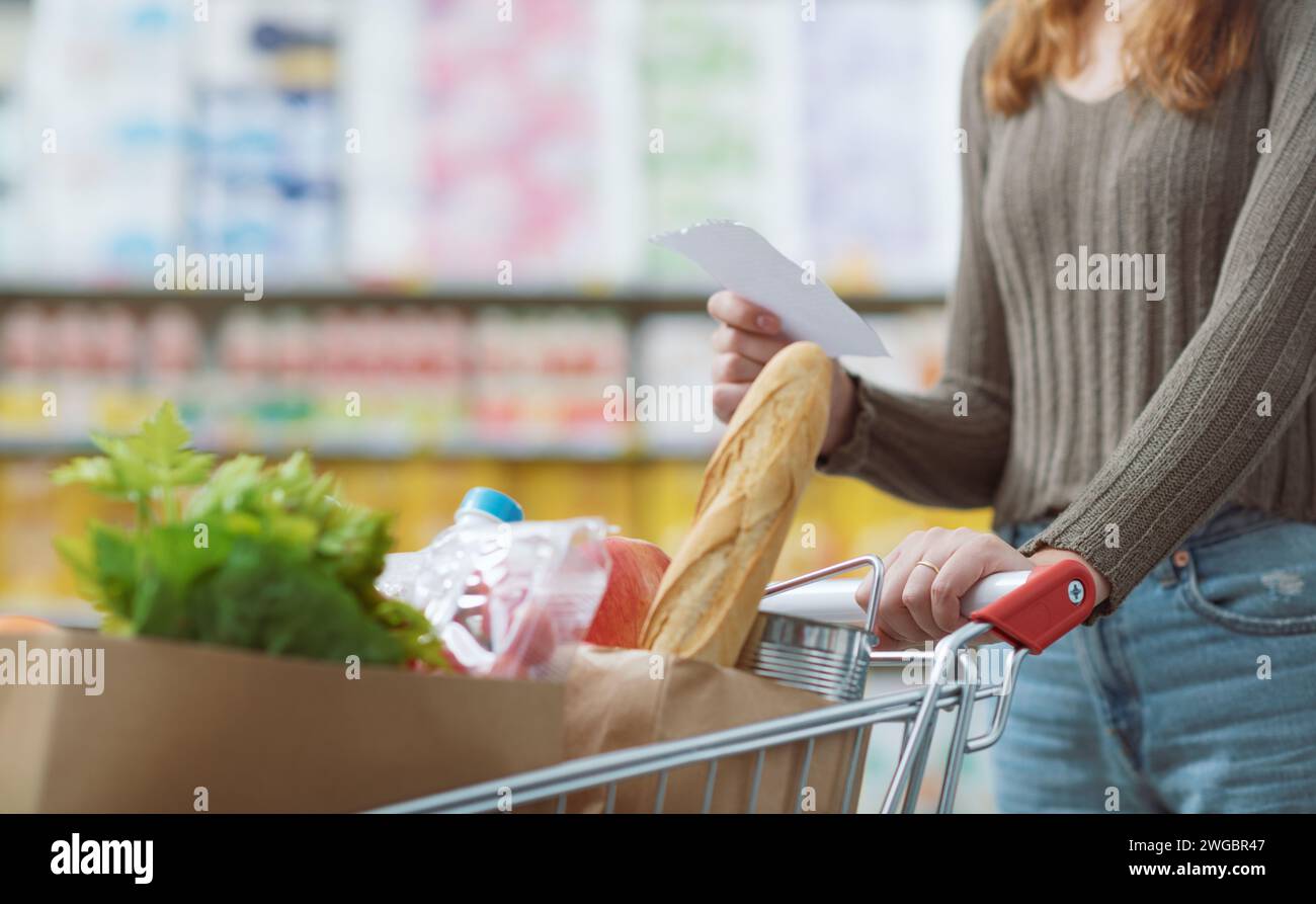 Eine junge Frau, die im Supermarkt einkaufen geht, schiebt einen vollen Trolley und hält eine Einkaufsliste, Nahaufnahme Stockfoto