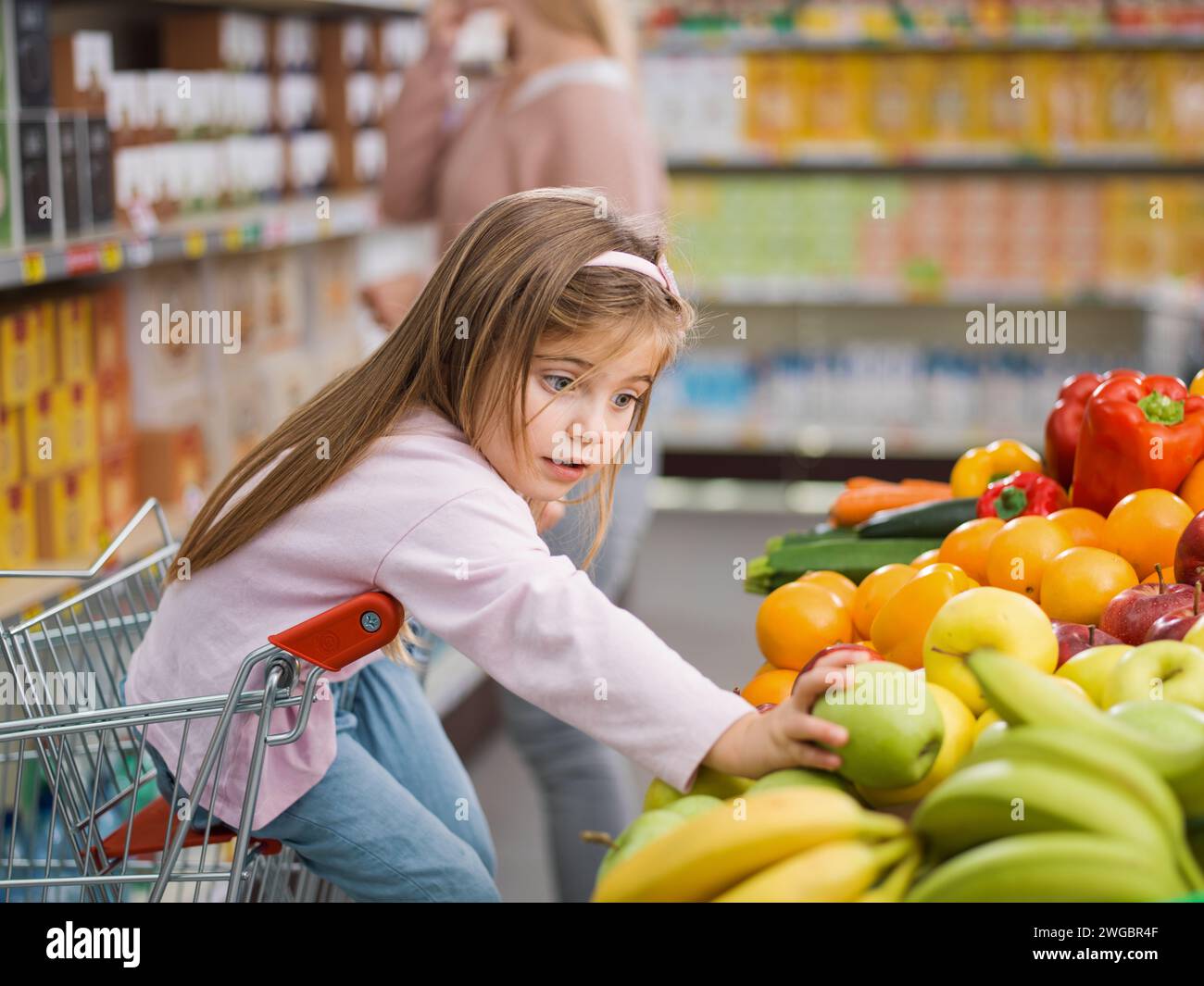 Niedliches, hübsches Mädchen, das auf dem Einkaufswagen sitzt, nimmt eine Frucht in der Produktabteilung, während ihre Mutter damit beschäftigt ist, Produkte in die Regale zu nehmen Stockfoto