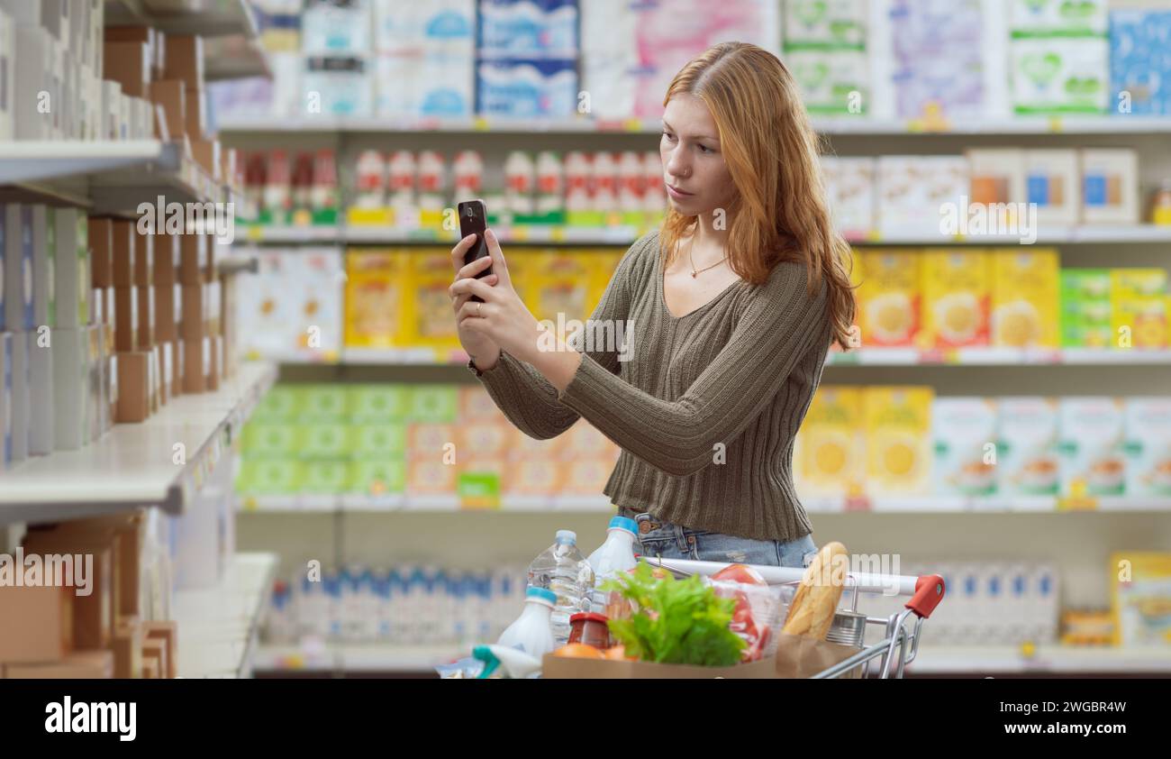 Eine junge Frau, die im Supermarkt Lebensmittel einkauft, überprüft Lebensmittelinformationen mithilfe einer Food Scanner-App auf ihrem Smartphone Stockfoto