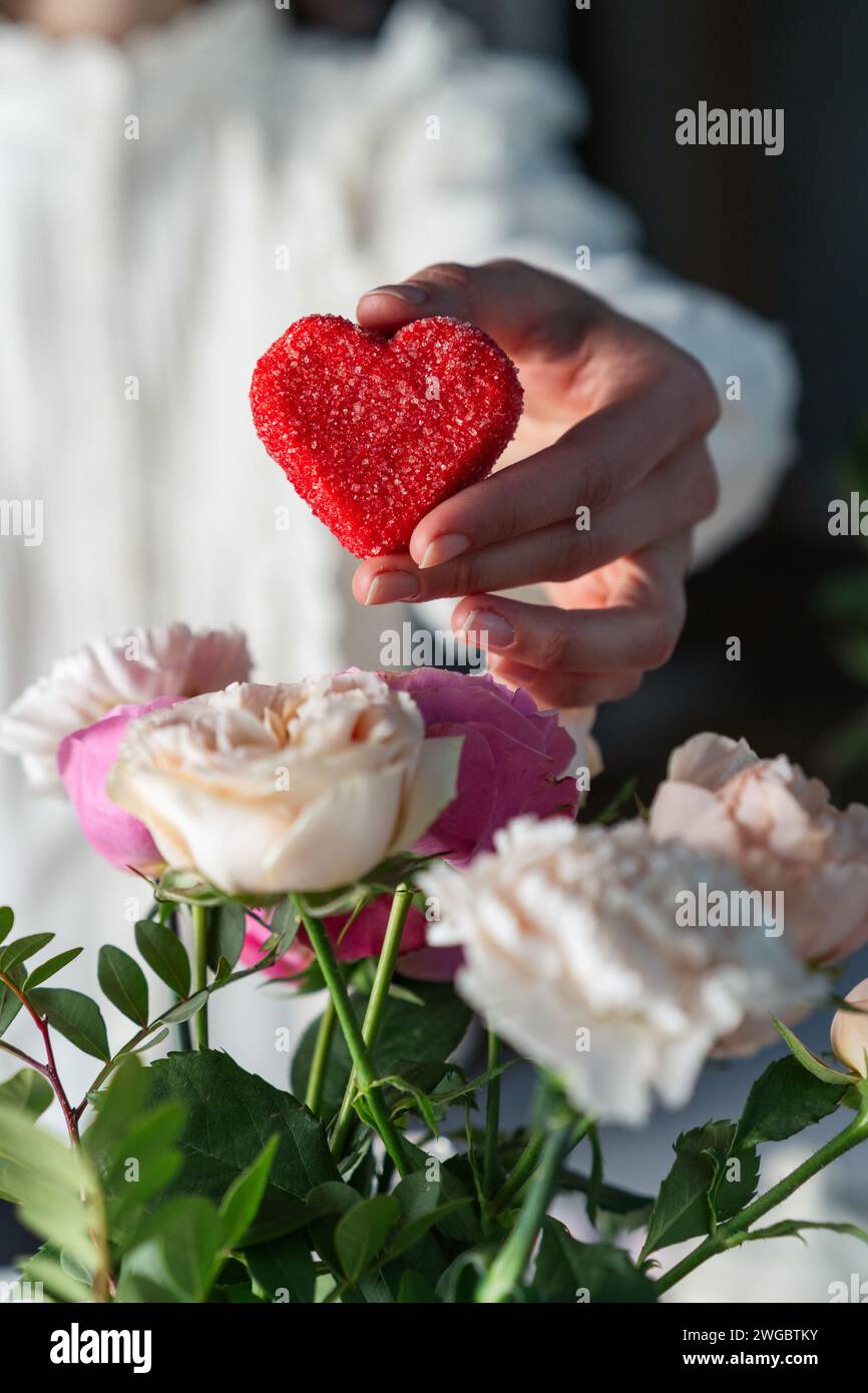 Die Hand der Frau hält eine zuckerbeschichtete rote Herzbonbons neben einem Blumenstrauß zum Valentinstag Stockfoto