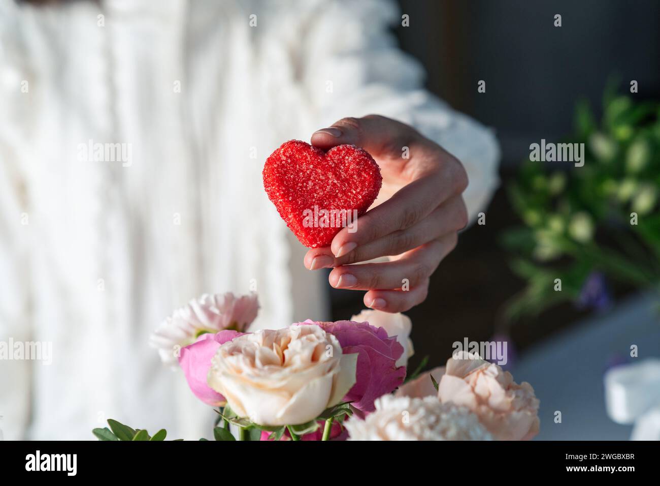 Die Hand der Frau hält eine zuckerbeschichtete rote Herzbonbons neben einem Blumenstrauß zum Valentinstag Stockfoto