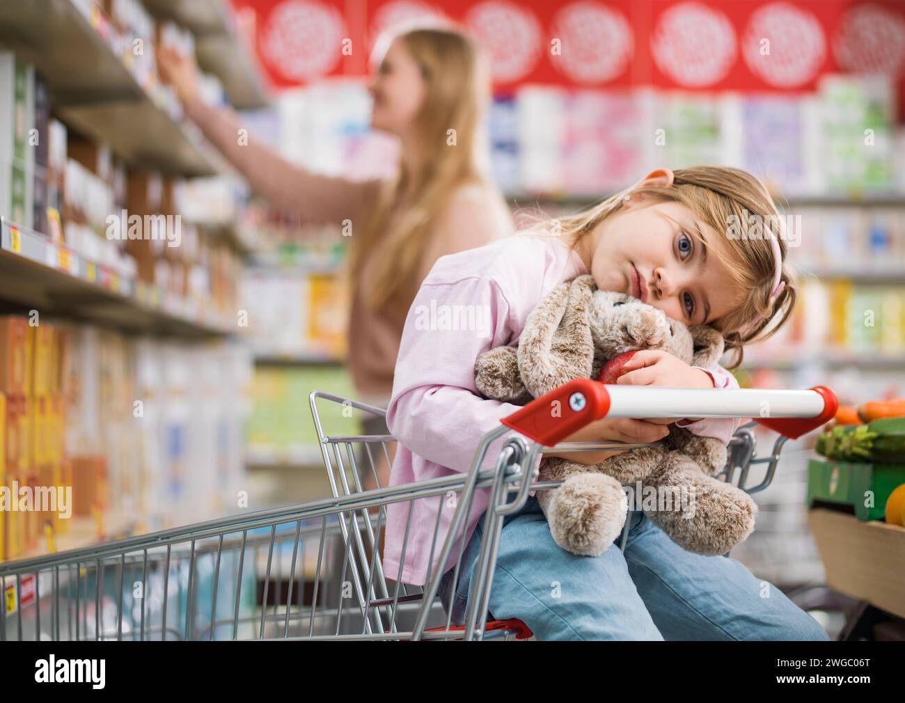 Niedliches gelangweiltes Kind, das auf einem Einkaufswagen sitzt und ihr Plüschtier umarmt, wartet auf ihre Mutter Stockfoto