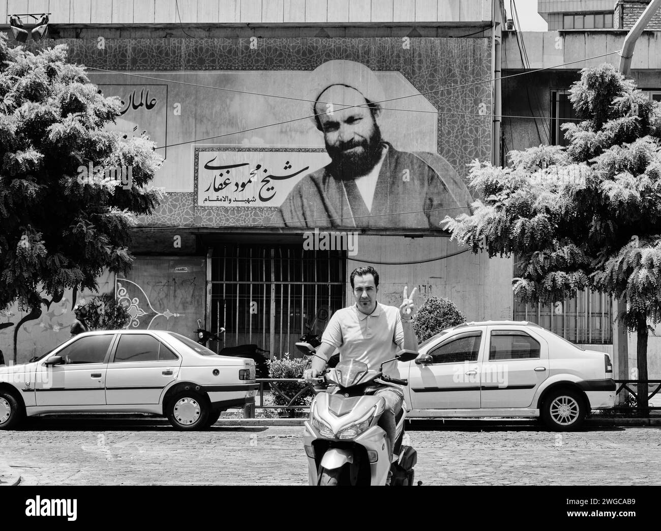 Teheran (Teheran), Iran, 06.24.2023: Ein iranischer Mann macht Siegeszeichen unter dem Plakat des Imams. Schwarzweißfoto des Iran. Stockfoto