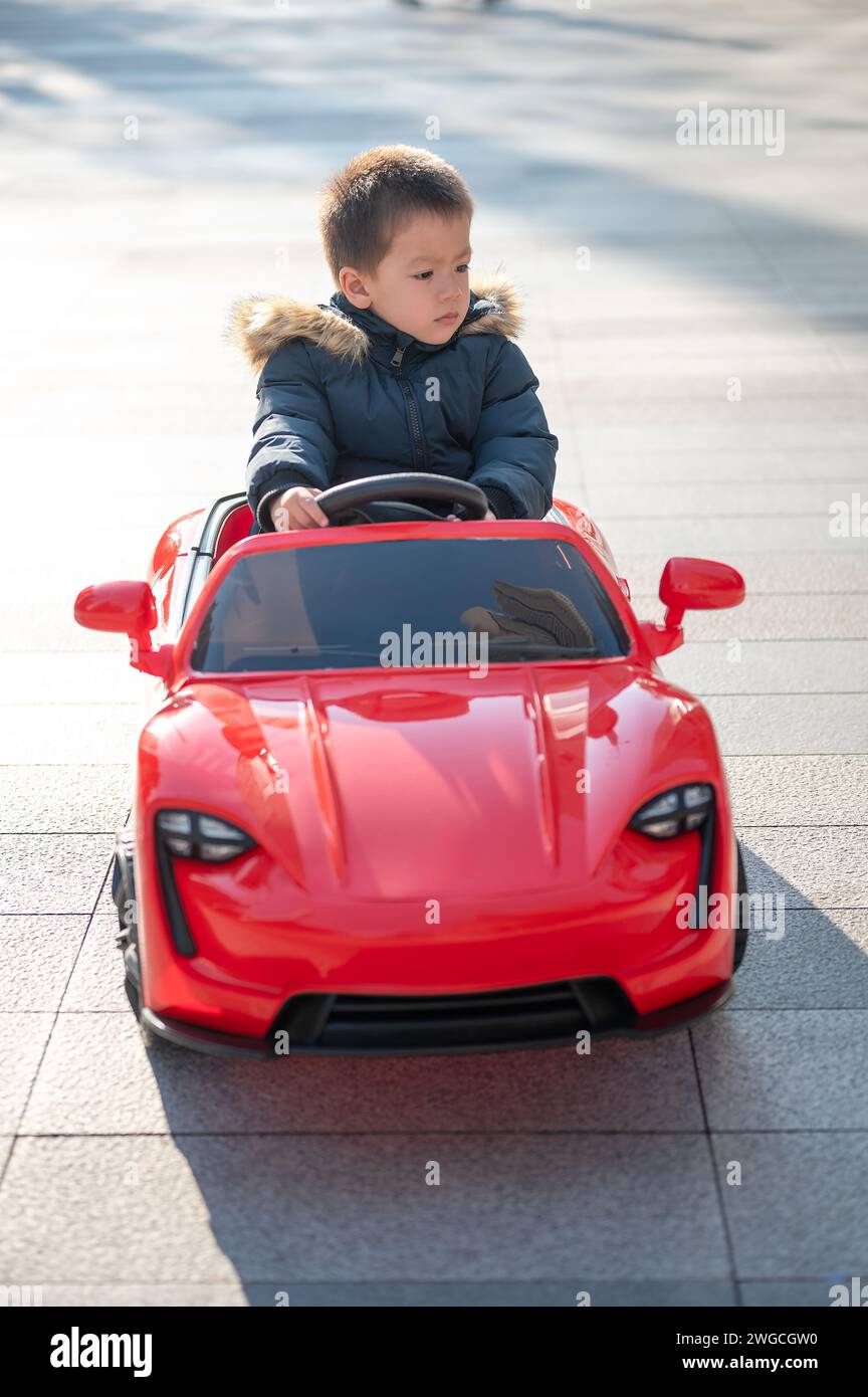 Ein junges, multirassisches Kleinkind navigiert freudig in einem roten Sportwagen über die Nachbarschaftsstraße und lässt ein großes Lächeln auf sich wirken, als es die Kontrolle über sein Spielzeug Ca übernimmt Stockfoto