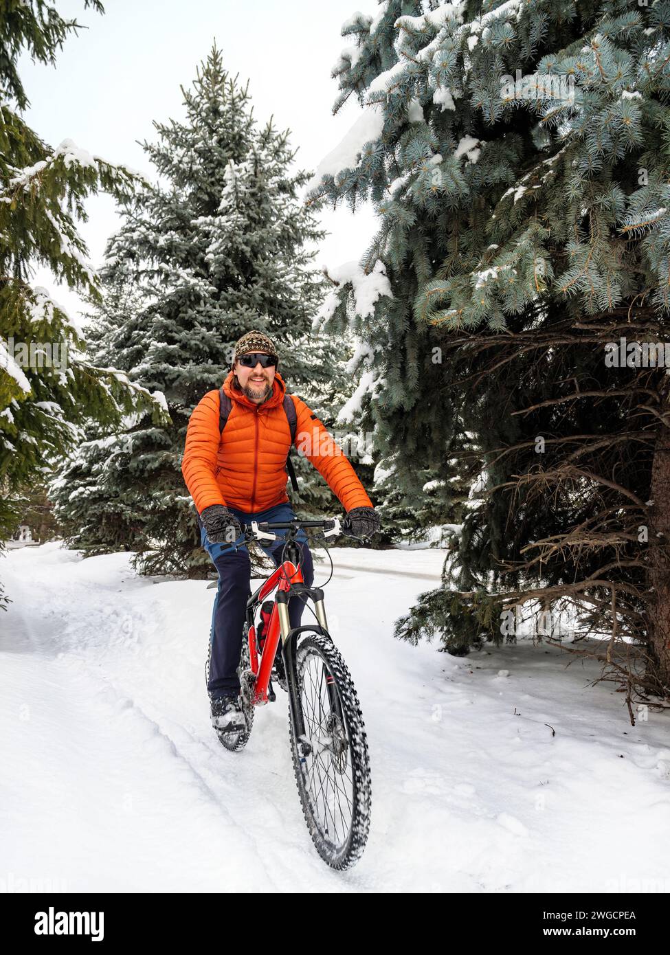 Ein Typ fährt mit dem Fahrrad in einem Winterpark. Ein Radfahrer in einer orangen Jacke fährt freudig auf einem verschneiten Pfad. Aktiver Lebensstil im Winter Stockfoto