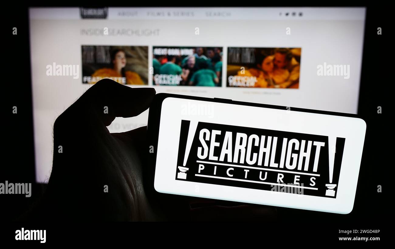Person mit Smartphone und Logo der US-Filmproduktionsfirma Searchlight Pictures vor der Website. Schwerpunkt auf dem Display des Telefons. Stockfoto