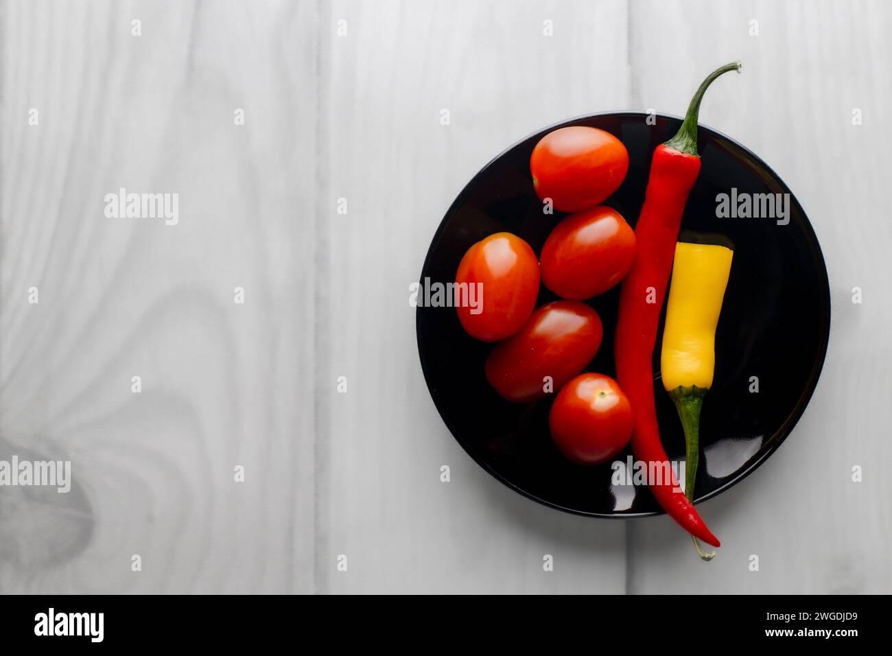 Anderthalb heiße Paprika auf einem schwarzen runden Teller. Kirschtomaten. Heller Hintergrund. Whiteboards. Gewürze in der Küche. Stockfoto