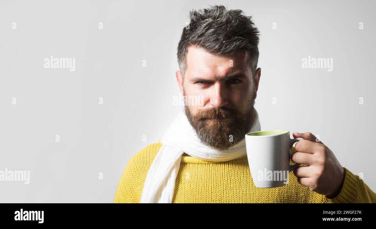 Ernster Mann in gestricktem Pullover in Schal mit einer Tasse Kaffee oder Tee gewickelt. Herbst- oder Winterzeit. Hübscher bärtiger Mann in warmer Kleidung, der trinkt Stockfoto