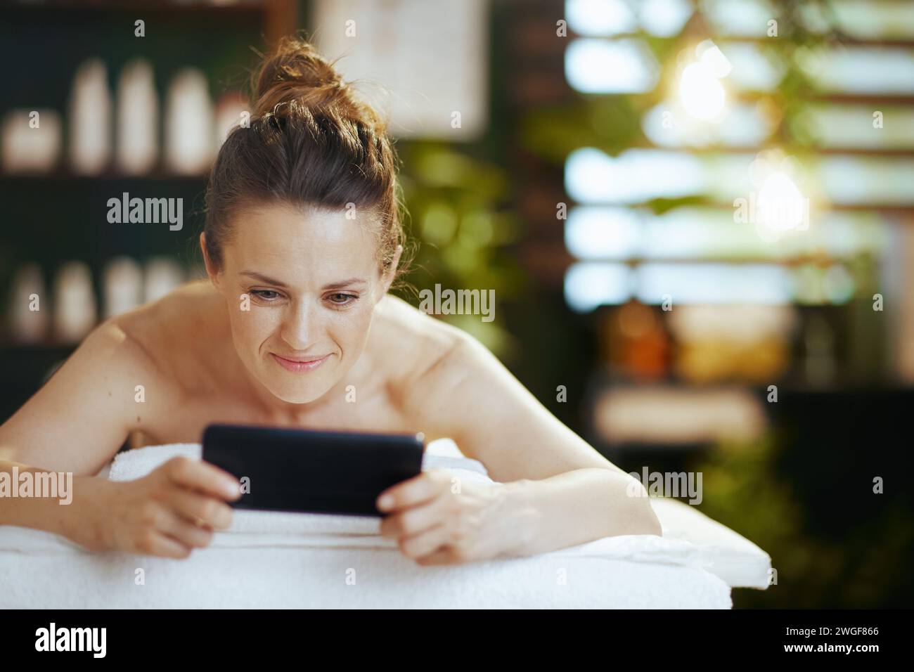 Gesundheitszeit. Entspannte, moderne Frau mittleren Alters im Massageschrank mit Smartphone und auf Massagetisch liegend. Stockfoto