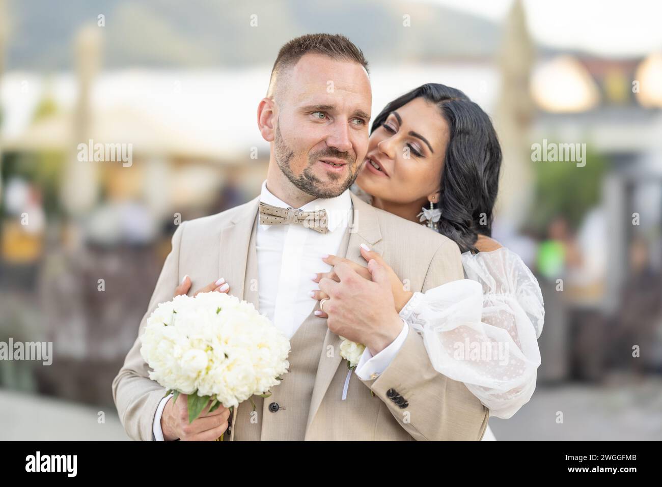 Hochzeitsporträt lächelnder Jungvermählter. Ein stilvoller Bräutigam in einem beigefarbenen Anzug und eine süße Brünette in einem weißen Kleid sind zärtlich im Park. Stockfoto