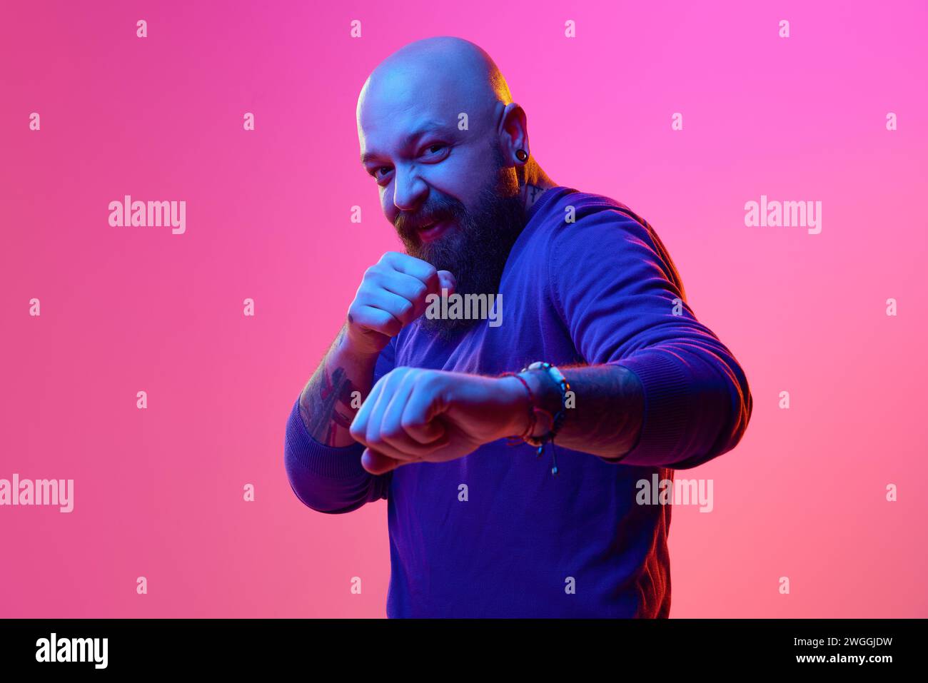 Lustiger Kampf. Gel-Porträt für künstlerischen bärtigen Glatzen-Mann in blauem Pullover, der sich emotional vor rosa Hintergrund im Neonlicht posiert Stockfoto