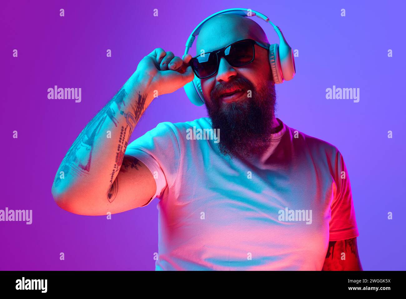 DJ. Bärtiger, künstlerischer Glatze Mann mit Tätowierungen, in weißem T-Shirt und Sonnenbrille, die vor verlaufendem lila Hintergrund im Neonlicht steht Stockfoto