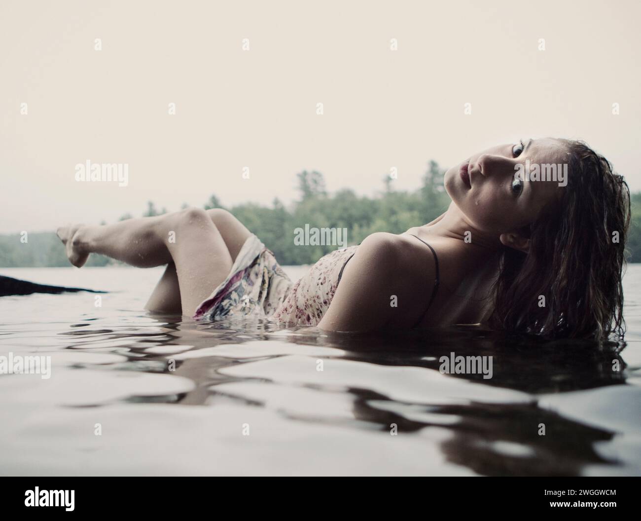 Eine junge Frau in einem weißen Kleid entspannt sich halb untergetaucht in einem See in Maine. Stockfoto