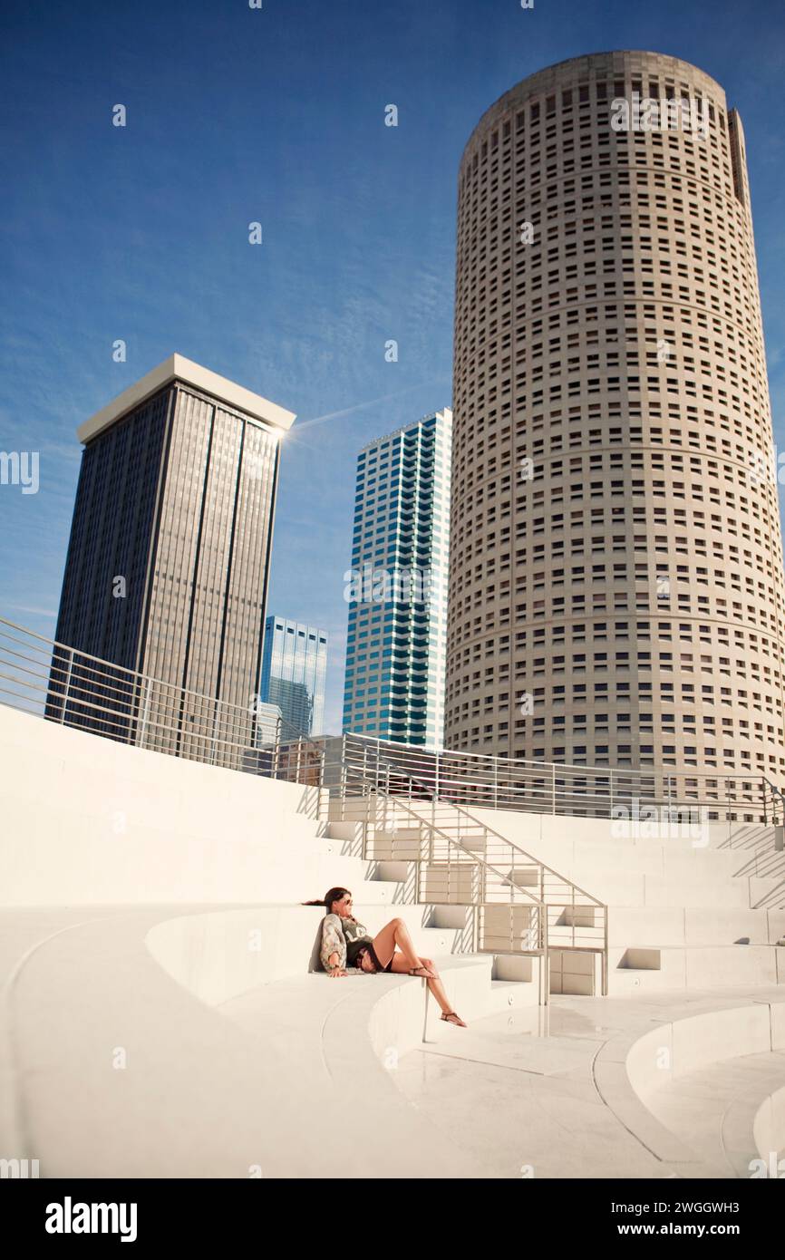 Eine Frau entspannt sich in der Sonne in einem Amphitheater, während sich die Wolkenkratzer der Skyline von Tampa, Florida, über ihr erheben. Stockfoto