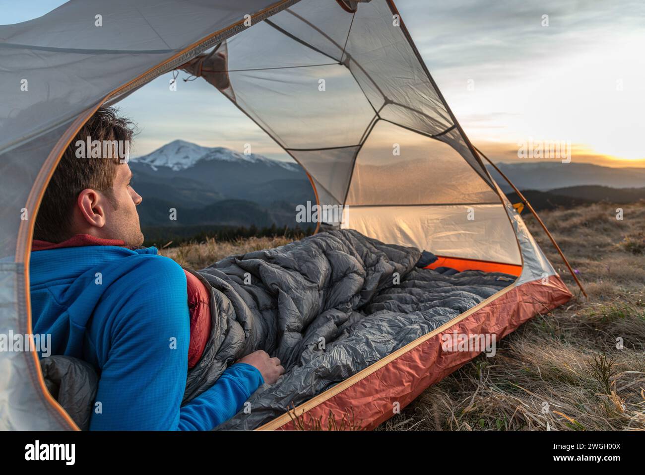 Junger Mann, der im Zelt ruht und den Sonnenuntergang in den Bergen sieht Stockfoto