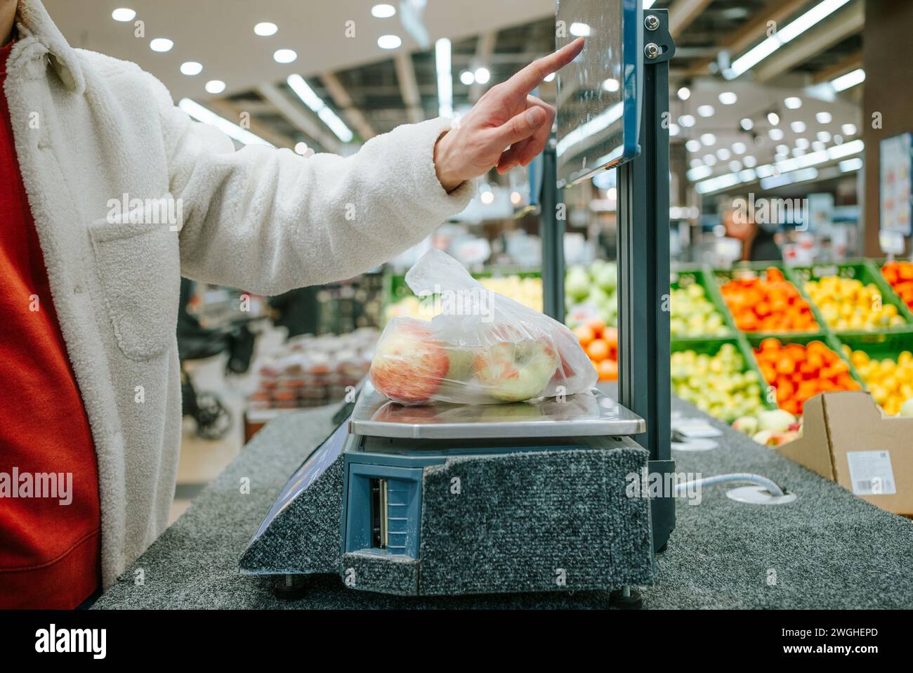 Der Finger des Mannes wählt das gewünschte Produkt im Supermarkt auf dem Touchscreen der elektronischen Waage aus. Person, die eine Tüte Äpfel an der Kasse eines Supermarktes wiegt. Stockfoto