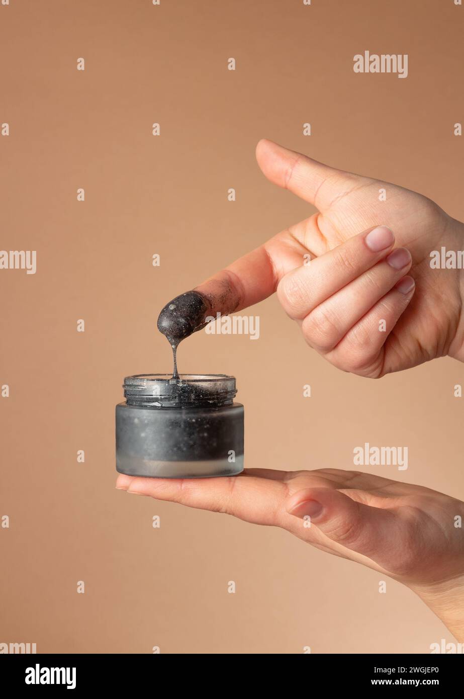 Weibliche Hände halten ein Glas mit schwarzer kosmetischer Gesichtspflege und tragen es auf den Finger auf braunem Hintergrund auf. Kosmetische Industrie. Stockfoto