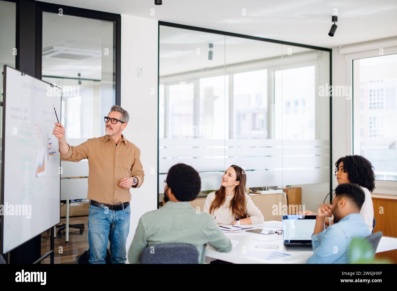 In einem hell beleuchteten Bürobereich führt ein reifer Mann mit grauen Haaren eine Diskussion vor einem Whiteboard und hebt wichtige Punkte mit einem Marker hervor, während sein vielfältiges Team von jungen Fachleuten aufmerksam zuhört Stockfoto