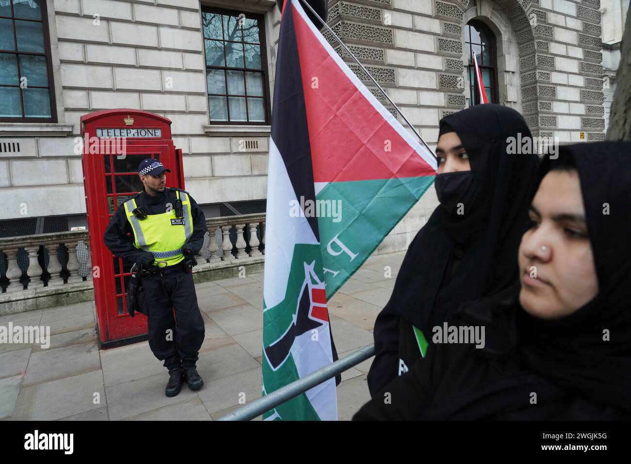 Ein Polizist steht vor einer roten Telefonzelle, während zwei muslimische Frauen bei einer pro-palästinensischen Kundgebung in Whitehall an einer palästinensischen Flagge stehen. Stockfoto