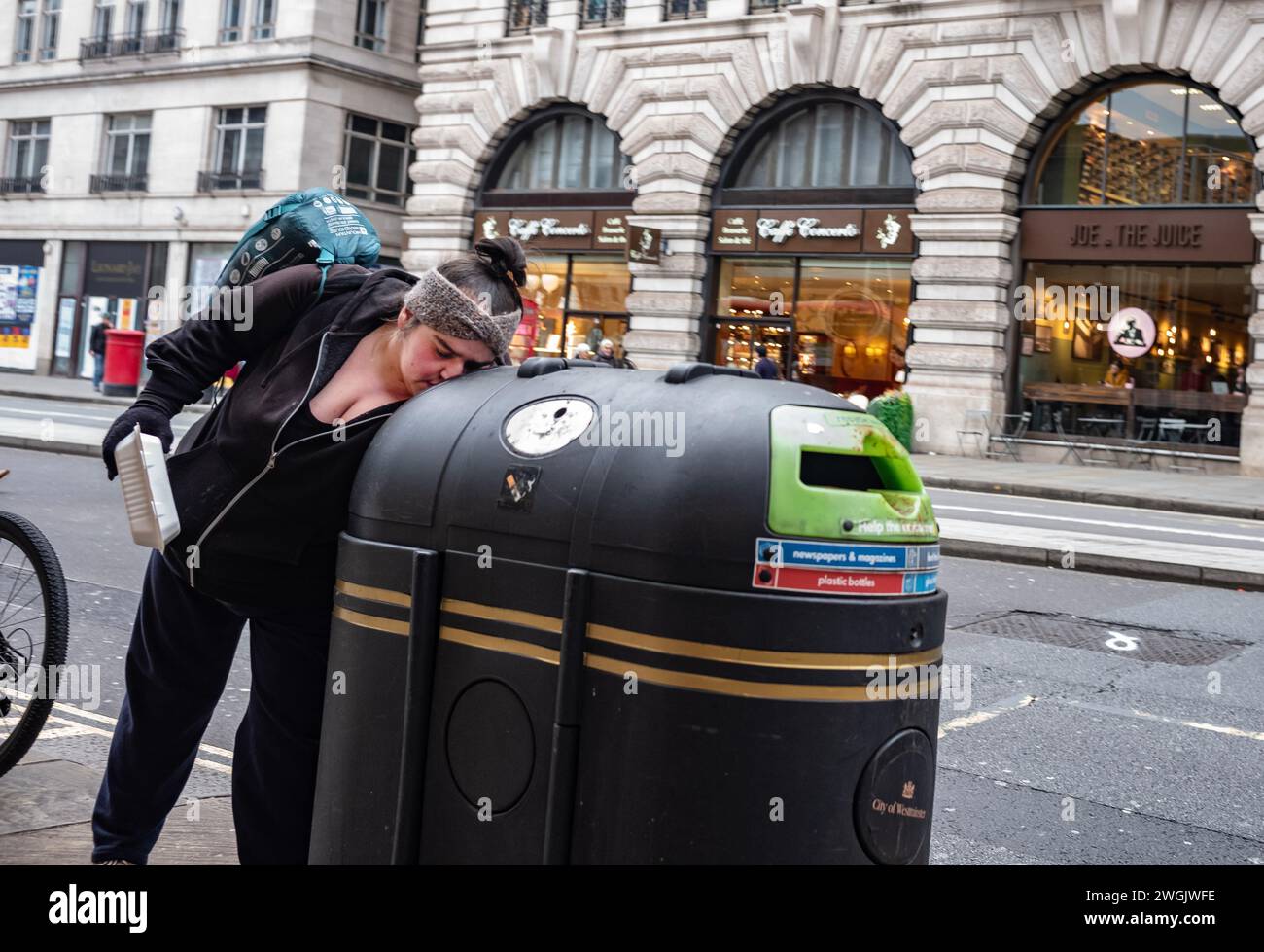 Obdachlose „Mülltauchen“ für ihren Tag Essen in einem öffentlichen Recyclingbehälter auf dem Bürgersteig entlang Piccadilly, Mayfair, London, England, Großbritannien Stockfoto