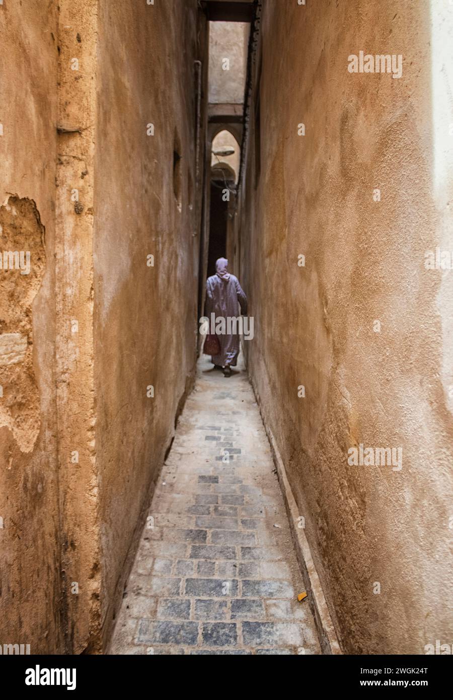 Ein Mann, der durch die engen Medina-Gassen von Fès läuft. Fès, die zweitgrößte Stadt Marokkos, wird oft als Kulturhauptstadt des Landes bezeichnet. Es ist vor allem bekannt für seine von Fes El Bali ummauerte Medina mit mittelalterlicher marinidischer Architektur, pulsierenden Souks und der Atmosphäre der alten Welt. Die Medina beherbergt religiöse Schulen wie die Bou Inania aus dem 14. Jahrhundert und Al Attarine, die beide mit aufwändigen Zedernschnitzereien und kunstvollen Fliesen verziert sind. Marokko. Stockfoto