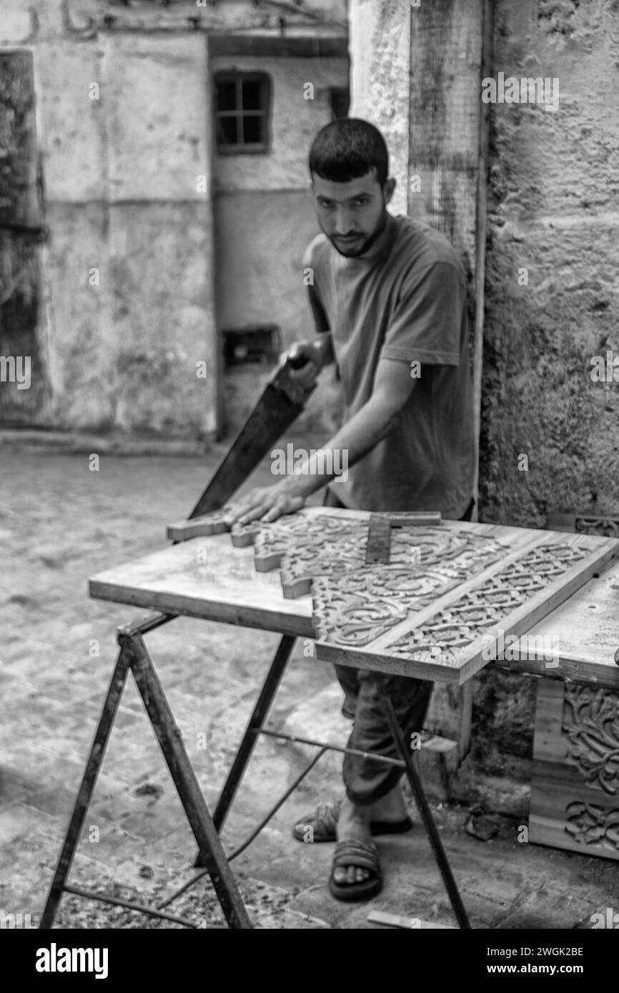 Ein handwerklicher Zimmermann, der in einer Medina-Straße in Fès Holzplatten herstellt. Fès, die zweitgrößte Stadt Marokkos, wird oft als Kulturhauptstadt des Landes bezeichnet. Es ist vor allem bekannt für seine von Fes El Bali ummauerte Medina mit mittelalterlicher marinidischer Architektur, pulsierenden Souks und der Atmosphäre der alten Welt. Die Medina beherbergt religiöse Schulen wie die Bou Inania aus dem 14. Jahrhundert und Al Attarine, die beide mit aufwändigen Zedernschnitzereien und kunstvollen Fliesen verziert sind. Marokko. Stockfoto